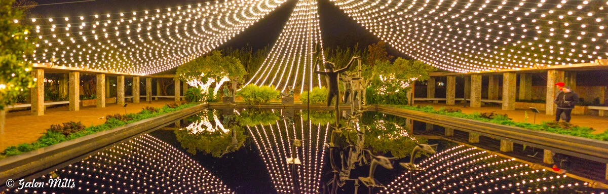 Outdoor pavilion with warm string lights reflecting in a pond, surrounded by greenery and statues at night.