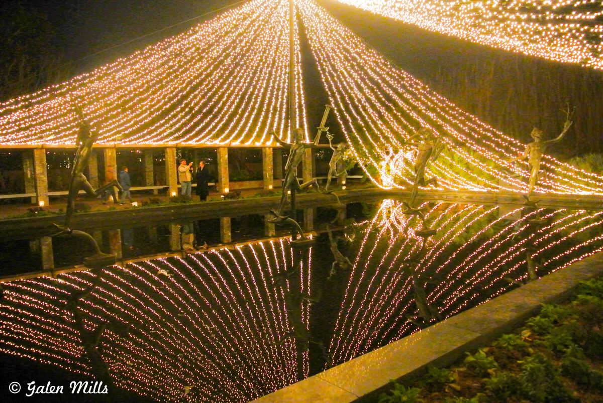 Night scene of outdoor lights hanging in a tent-like form over a reflecting pool with statues visible and people standing nearby.