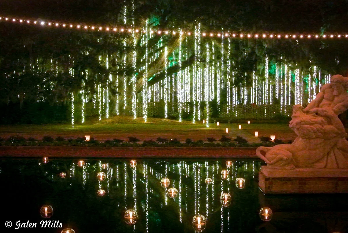Night scene with a string of lights hanging from trees, pond with floating candles, and a stone statue in the foreground.