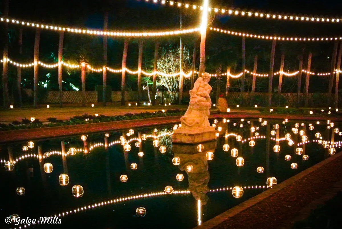 Outdoor fountain with stone statue surrounded by string lights and reflections in water at night.