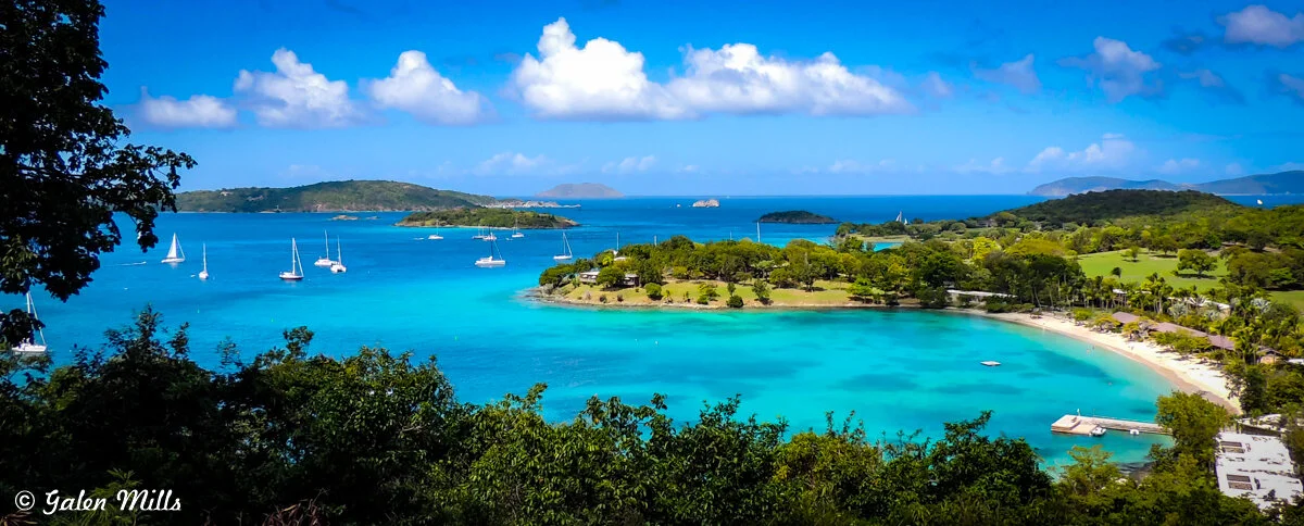 Scenic view of a tropical beach with turquoise water, lush green vegetation, and sailboats anchored. The horizon features small islands under a clear blue sky with fluffy clouds.