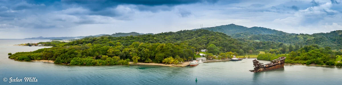 Panoramic view of a lush, green island with dense forest and a few scattered buildings. A rusted shipwreck is visible in the water near the shore. The sky is cloudy, suggesting an overcast day.