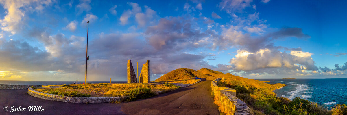 Panoramic view of a coastal landscape at sunset, featuring an asphalt road leading to triangular stone structures, an ocean, clouds, and hills.