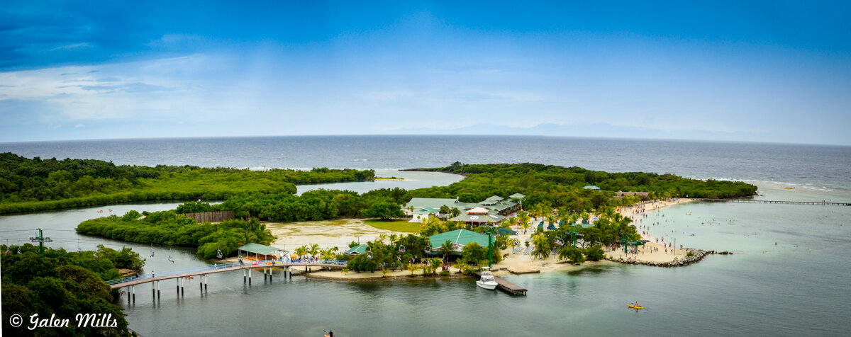 Aerial view of a tropical island with lush greenery, sandy beaches, and a wooden dock extending into the water. People are enjoying the beachfront area. The sea surrounds the island, with clear blue skies above.