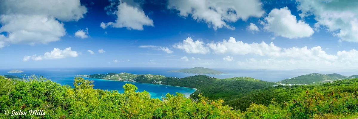 Panoramic view of lush green hills, a bright blue ocean, and scattered clouds in a clear sky.