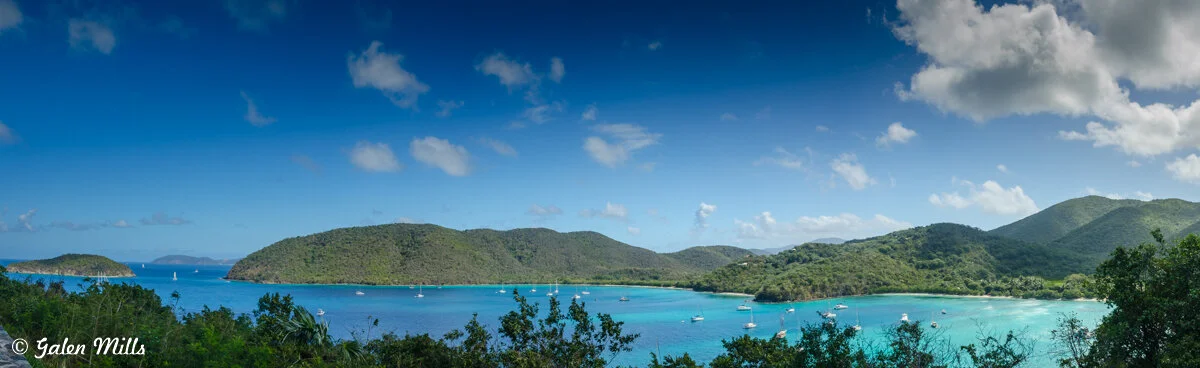 Panoramic view of a tropical bay with turquoise water and lush green hills in the background; boats are scattered across the water, and trees frame the scene.
