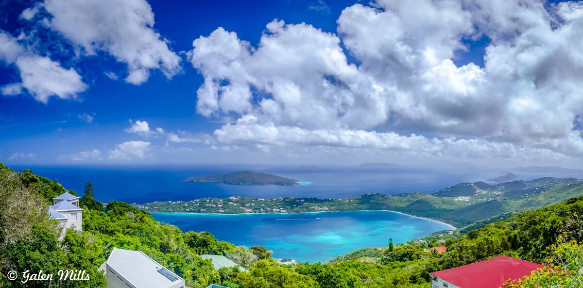 Panoramic view of lush green hills overlooking a blue ocean with islands in the distance under a partly cloudy sky.