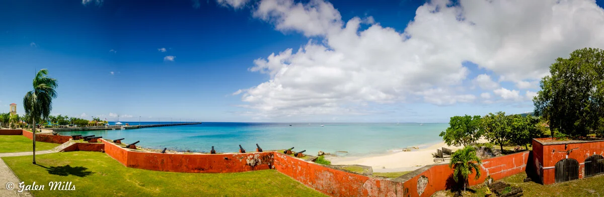 Panoramic view of a beach with turquoise water, sandy shore, red brick fort walls, palm trees, and a pier in the distance. The sky is partly cloudy.