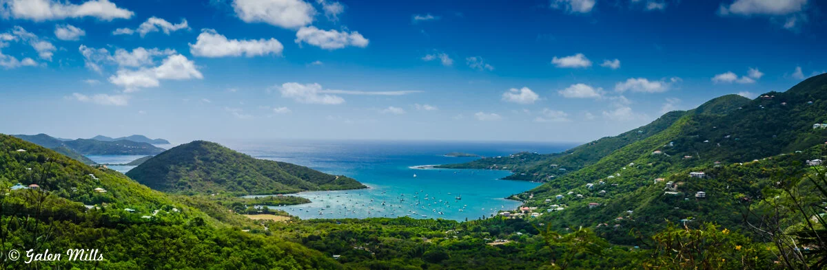 Scenic view of a coastal landscape with green hills and numerous boats in a bright blue bay under a cloudy sky.