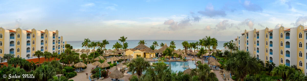Panoramic view of a tropical resort featuring palm trees, thatched umbrellas, and a large central swimming pool flanked by two rows of beige buildings, facing the ocean.