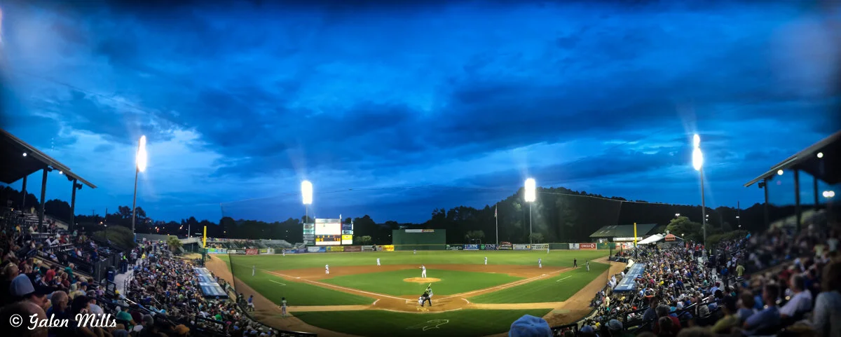 Panoramic view of a well-lit baseball stadium during a game, with an evening sky and crowd in the stands.