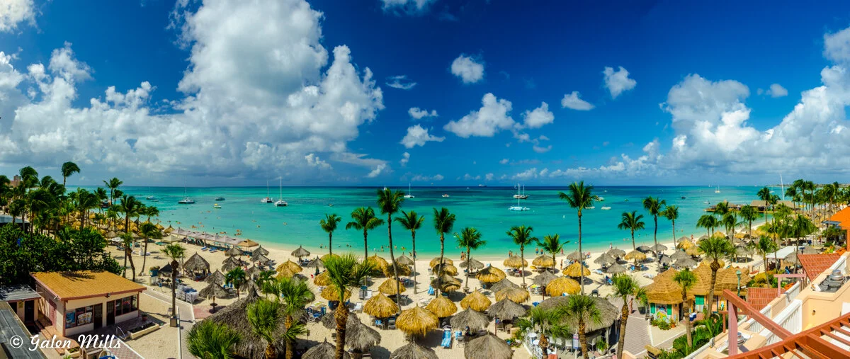 Panoramic view of a tropical beach with turquoise water, palm trees, and straw umbrellas, under a bright blue sky with scattered clouds. Boats are anchored near the shore.