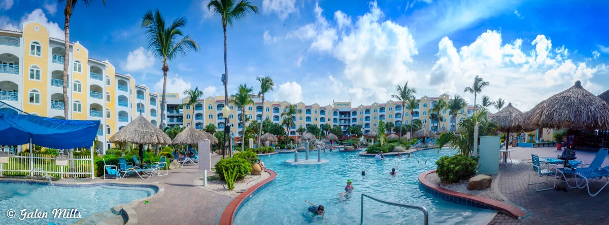 Resort pool area with palm trees and thatched umbrellas, surrounded by colorful buildings under a blue sky with clouds.