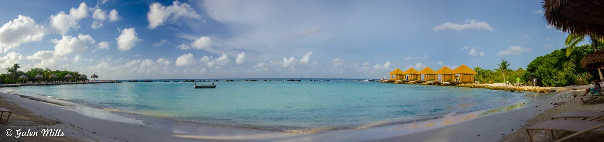 Panoramic view of tropical beach with turquoise water, thatched huts, and beach chairs under palm trees. Yellow bungalows line the shore under a blue sky with scattered clouds.
