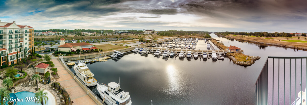 Panoramic view of a marina with numerous yachts, surrounded by buildings and greenery under a cloudy sky.