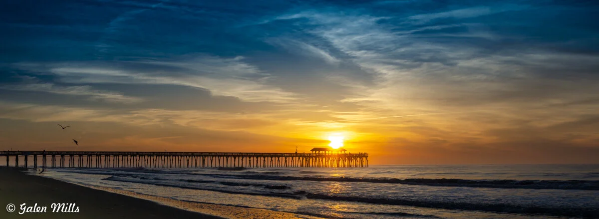Scenic ocean view with a wooden pier at sunset, waves gently crashing on the shore, and a colorful sky.