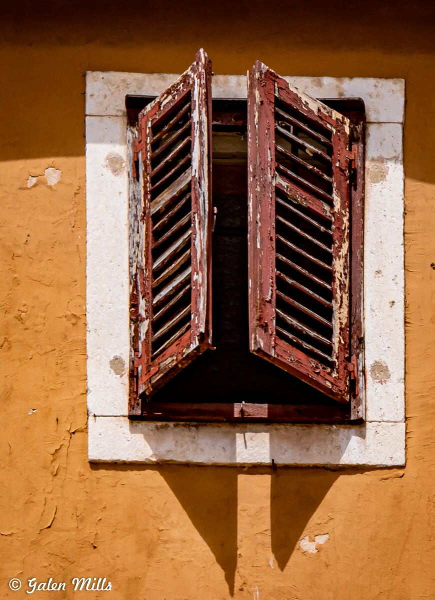 Weathered red wooden shutters on a rustic orange wall with a white stone frame.