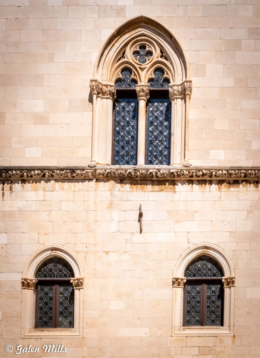 Architectural facade with three Gothic-style arched windows featuring circular glass patterns and decorative stone carvings.