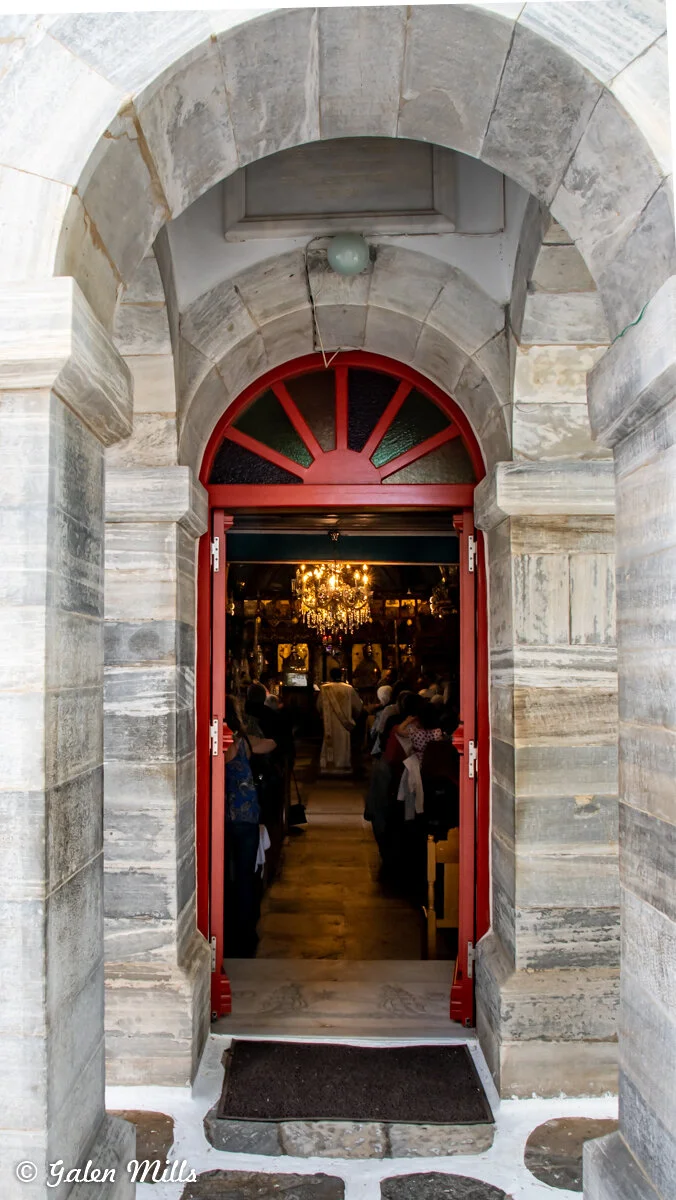 Arched stone doorway with red-framed entrance leading into a dimly lit interior with a chandelier and people seated inside.