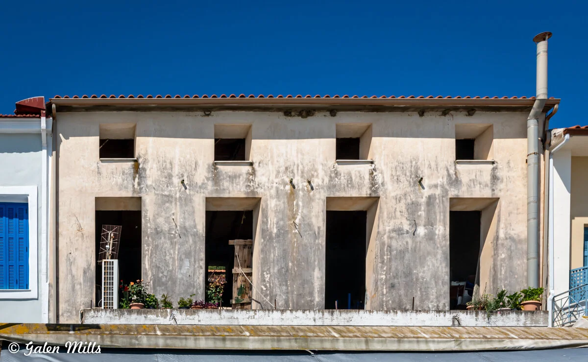 Old building with weathered façade, rectangular openings, and small balcony with plants against a blue sky.