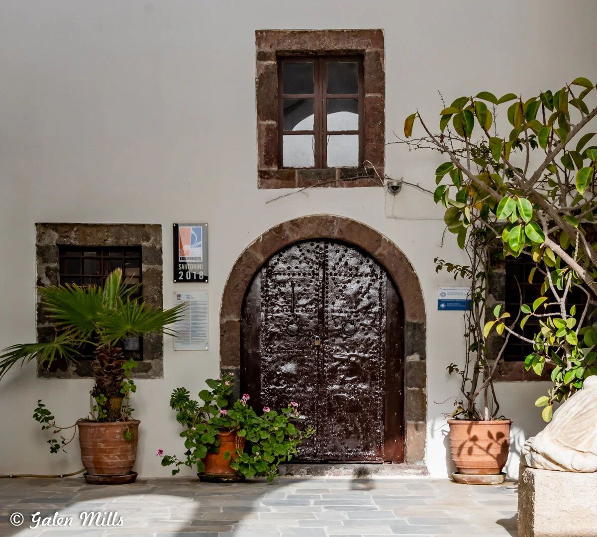 A traditional Greek building entrance featuring a wooden arched door with metal studs, surrounded by a stone frame. Above the door is a small window, and to the sides are potted plants including a palm and a leafy shrub. The building is labeled with 