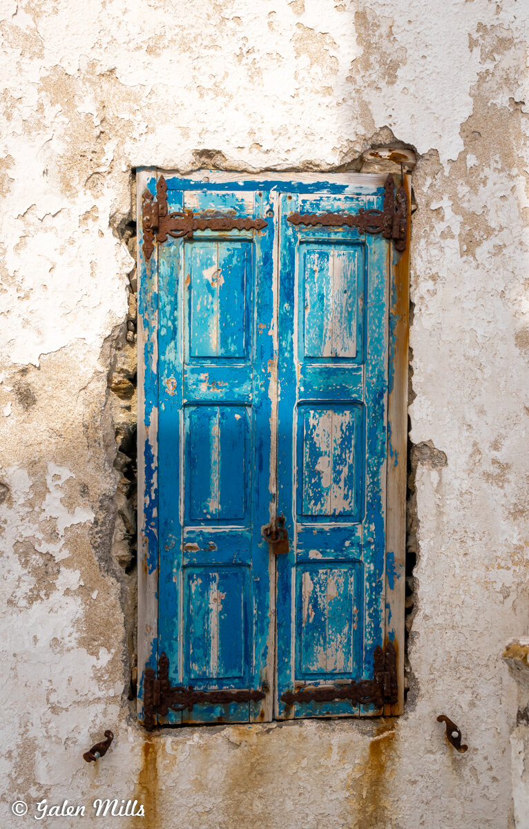 A weathered blue window shutter with peeling paint on a textured white wall, featuring rusted hinges and locks.