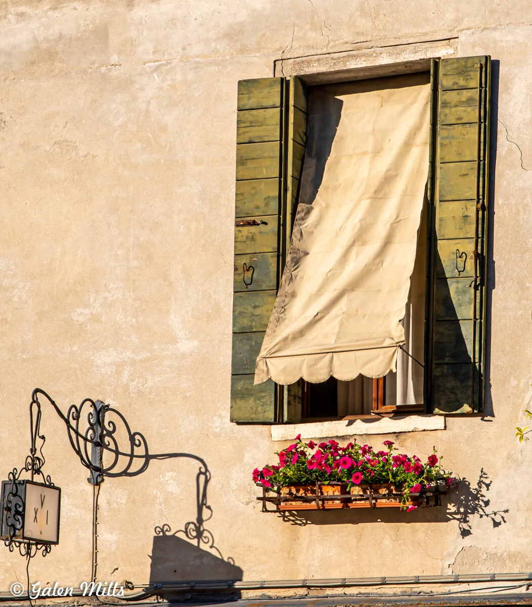 Old building exterior with a window featuring green shutters and a beige awning, accompanied by a flower box filled with pink flowers. An ornate metal sign hangs nearby casting a shadow.