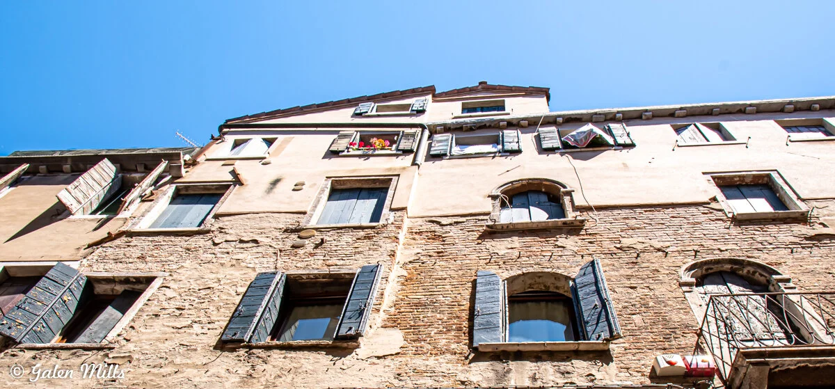 Low-angle view of a historic building with weathered stone and brick facade, featuring blue wooden shutters and flower boxes, under a clear blue sky.