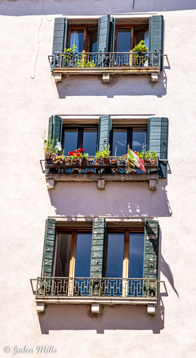 View of three stacked balconies with green shutters and potted plants on a light-colored building wall. Each balcony features decorative railings, and one has a small flag.