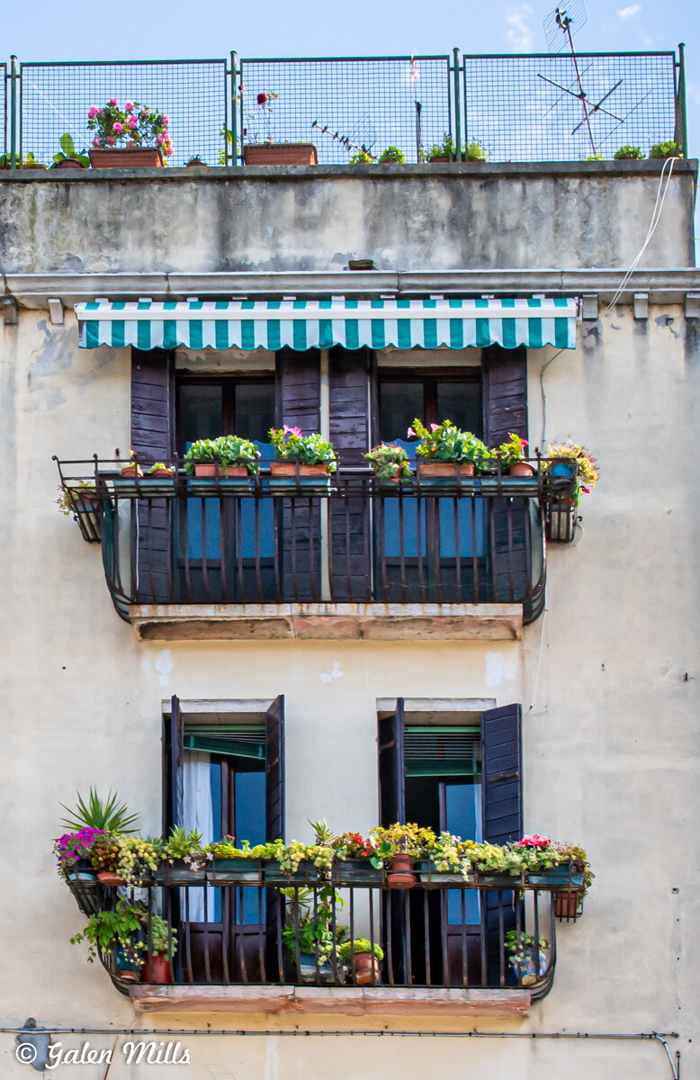 Two balconies with potted plants and flowers on an old building facade. The upper balcony has a green and white striped awning, while the lower one displays more greenery. Visible shutters on windows.