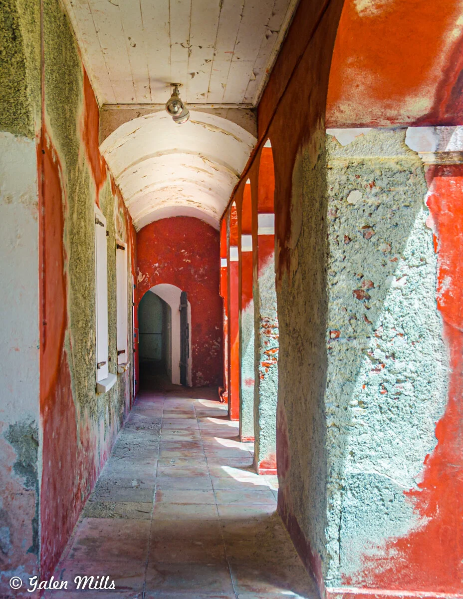 Narrow rustic corridor with red and beige textured walls, arched openings, and a tiled floor, leading to a shadowy doorway.
