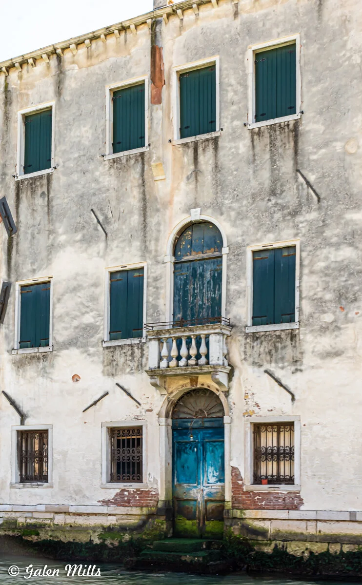Old building with closed shutters and peeling paint, featuring a blue door and small balcony. The facade shows signs of aging and weathering.