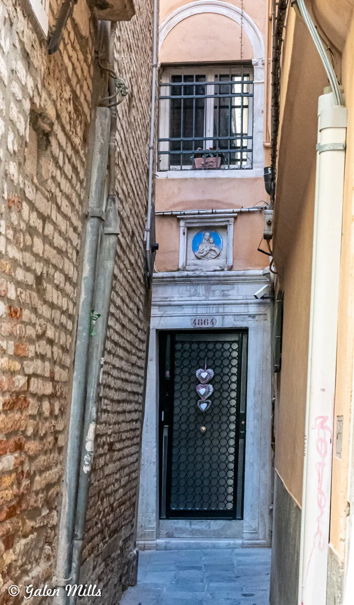 Narrow alleyway with a view of a doorway featuring heart decorations and an adjacent building with a high window and religious icon in relief above the door.