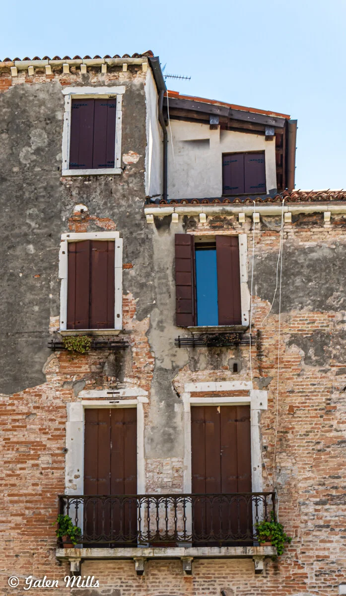 Old brick building with closed wooden shutters and small balcony. Weathered facade and climbing plants visible.