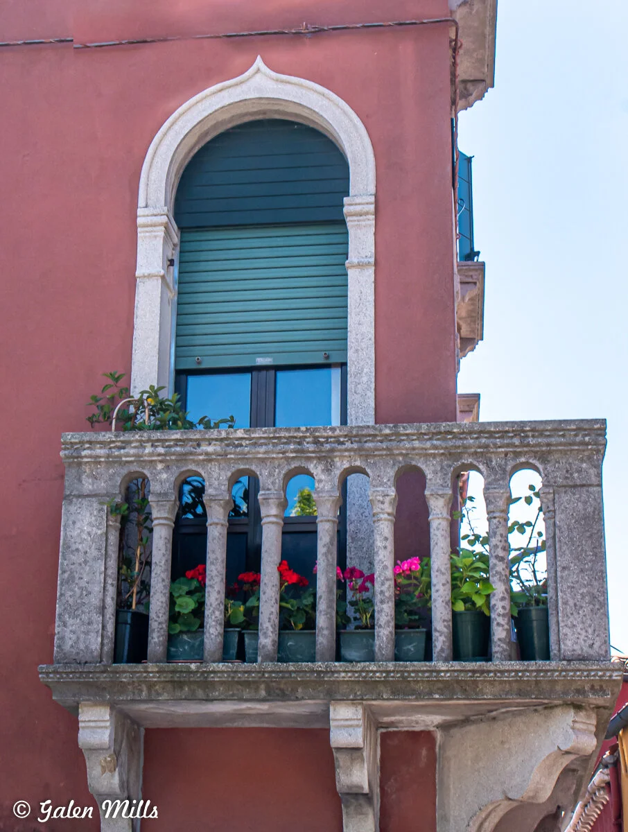 A pink stucco building featuring a small stone balcony adorned with potted flowers, showcasing colorful blooms. The window behind has an arched stone frame and closed wooden shutters.