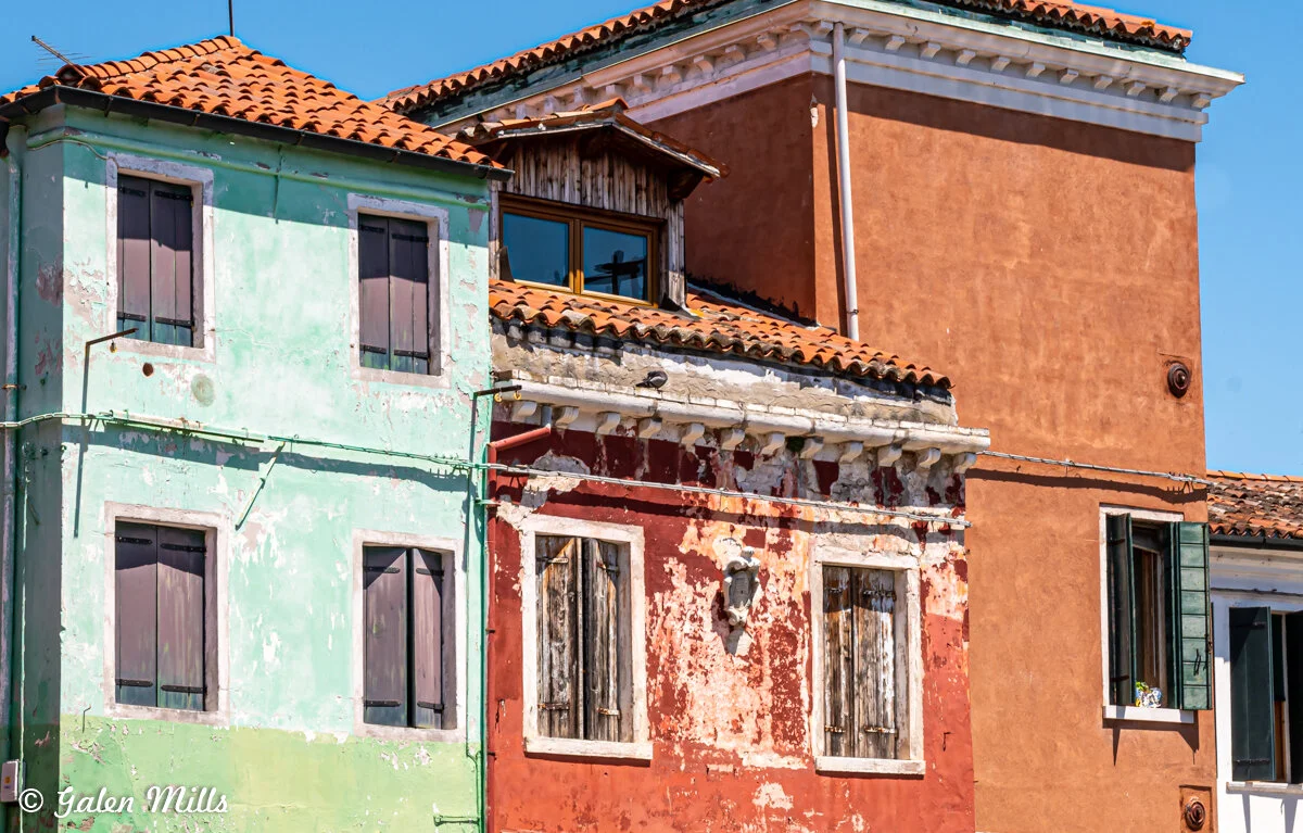 Colorful weathered buildings with peeling paint and closed wooden shutters, showcasing reddish, greenish, and brown facades, under a clear blue sky.