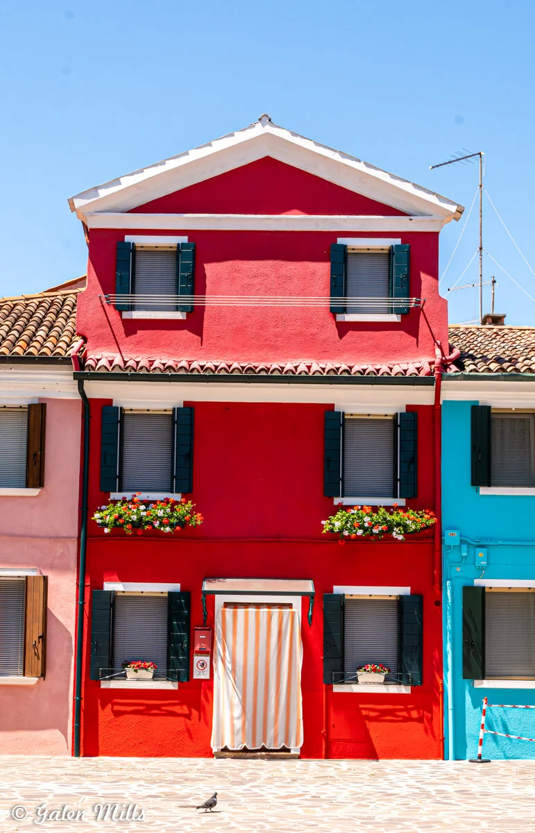 Colorful red house with white trim, green shutters, and flower boxes; adjacent to pink and blue buildings.