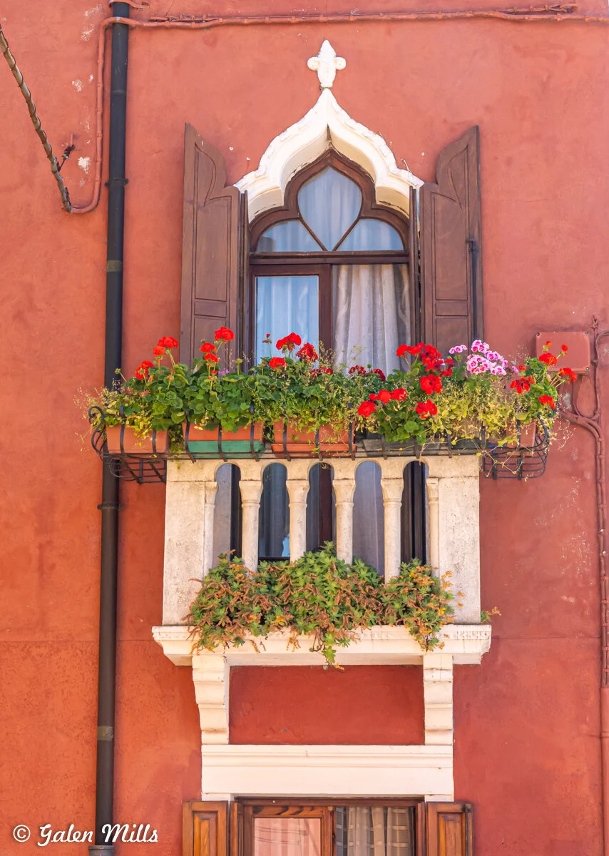 Ornate arched window with wooden shutters and a small balcony adorned with red and pink flowers against a red stucco wall.