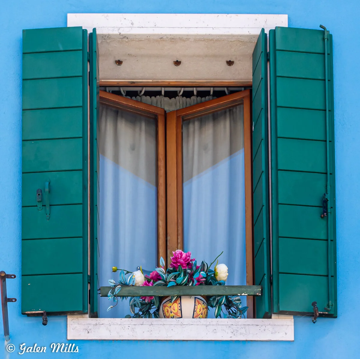 A blue wall with a window framed in stone, featuring green shutters and light curtains. Below the window, a decorative pot holds pink and white flowers with green leaves.