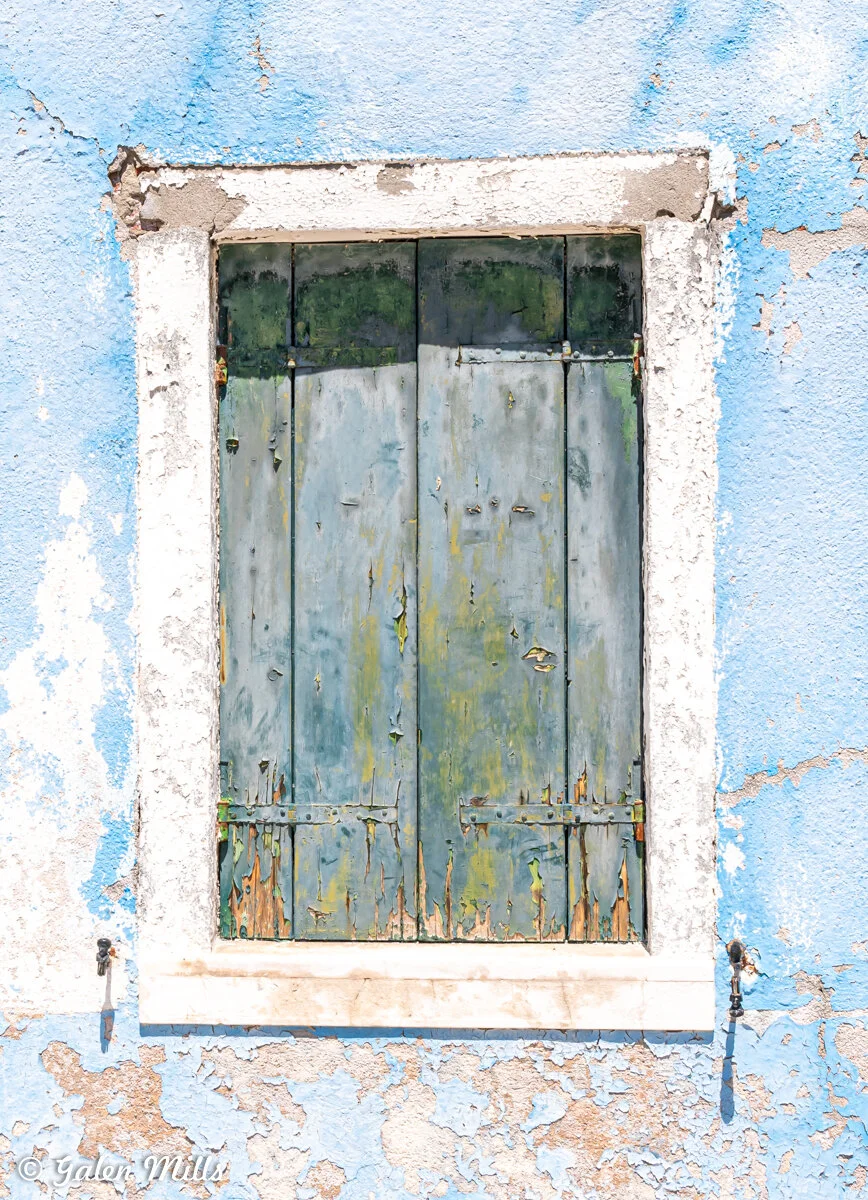 Rustic wooden shutters on a weathered wall with peeling blue paint and white trim.