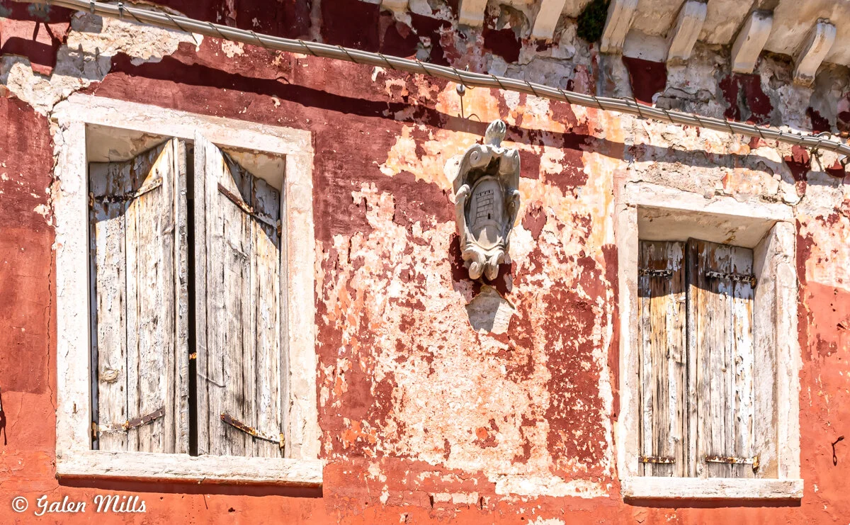 Two weathered wooden windows with shutters on a red, peeling plaster wall, featuring a decorative white stone emblem in between.