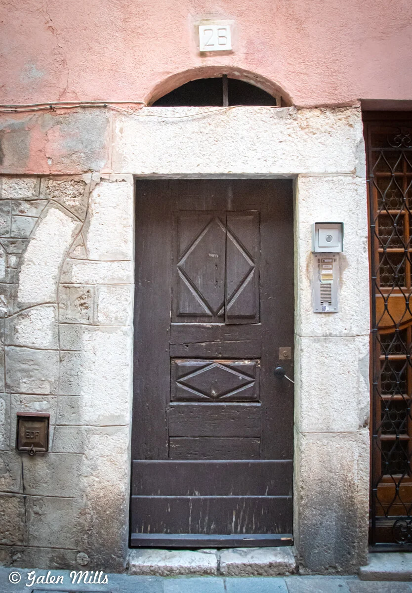 A weathered wooden door with decorative diamond patterns on an old stone building. Above the door is a plaque with the number 28, and to the right is an intercom system. The building has a rustic, aged appearance with a mix of stone and plaster.