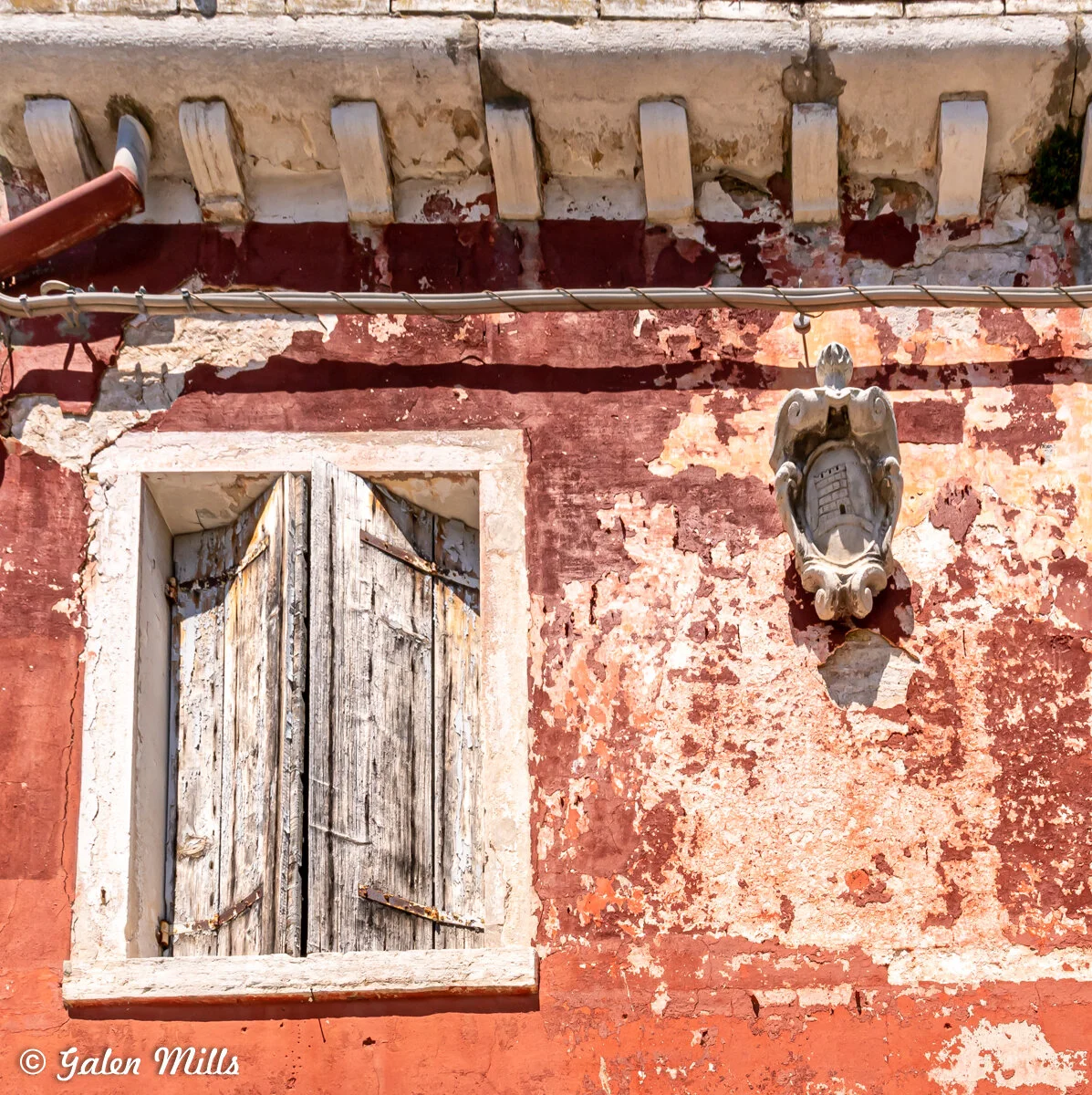 Weathered red wall with a worn wooden window shutter and ornate stone crest, showing signs of peeling paint and decay.