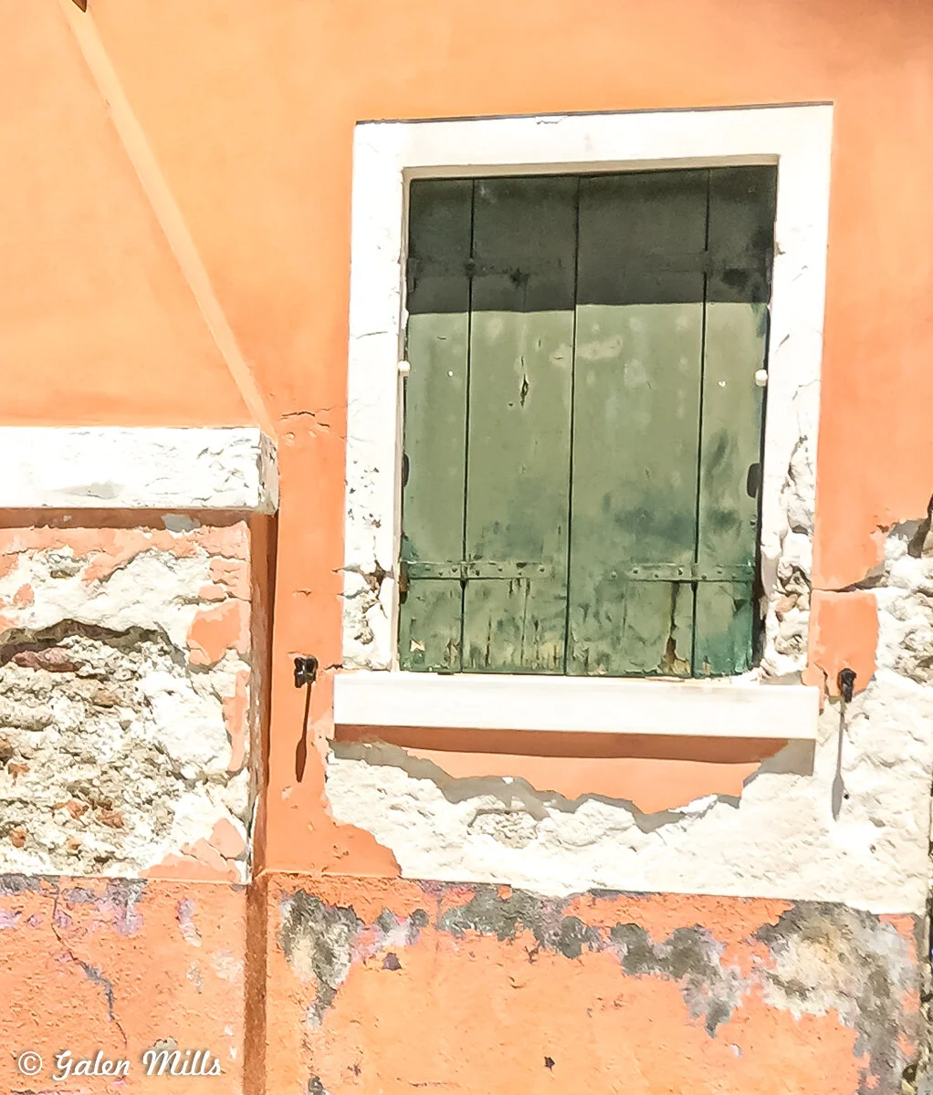 Weathered building facade with green shuttered window, peeling orange paint, and exposed stone.