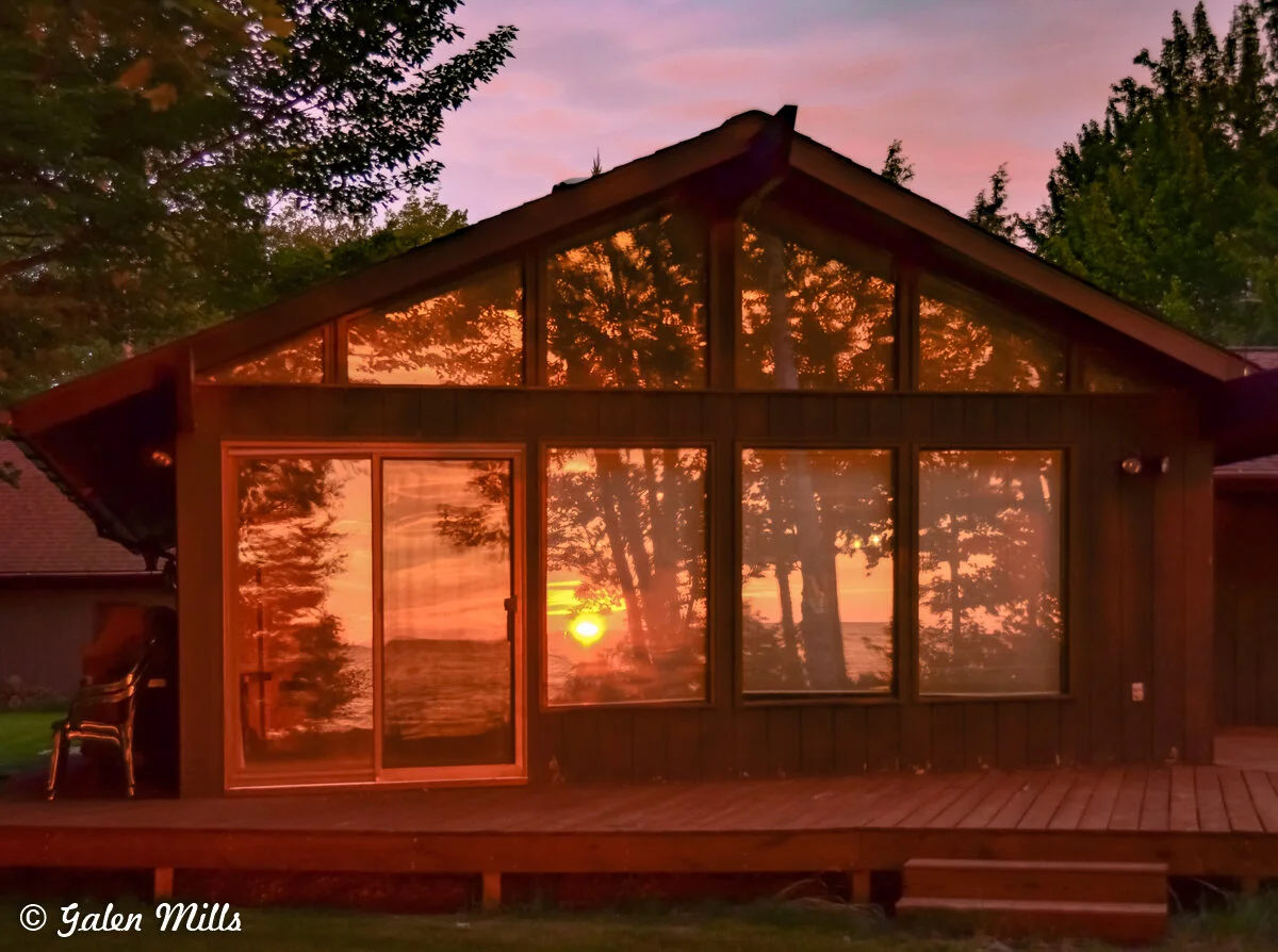 Reflection of a sunset in cabin windows, surrounded by trees.