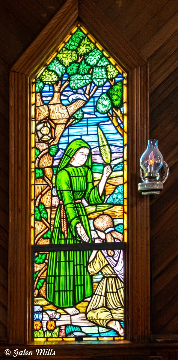 Stained glass window depicting a woman in a green robe holding a palm leaf, with a child kneeling beside her, surrounded by trees and flowers. A small lantern hangs on a tree branch. An oil lamp is mounted on the wall beside the window.