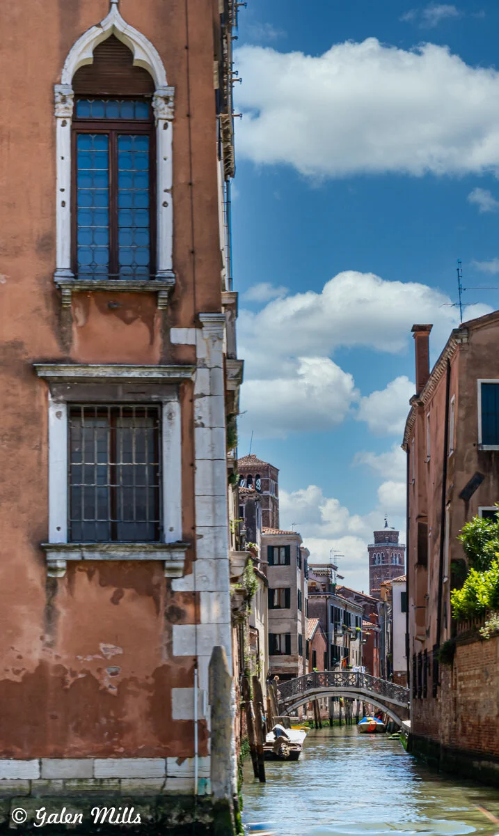Venetian canal scene with rustic buildings and a stone bridge under a blue sky.