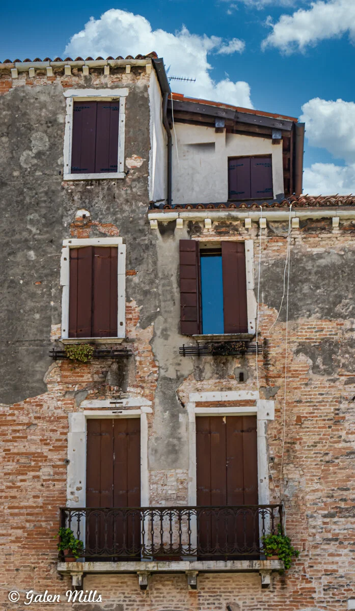 Old brick building facade with wooden shutters, small balcony, and climbing plants on a sunny day.
