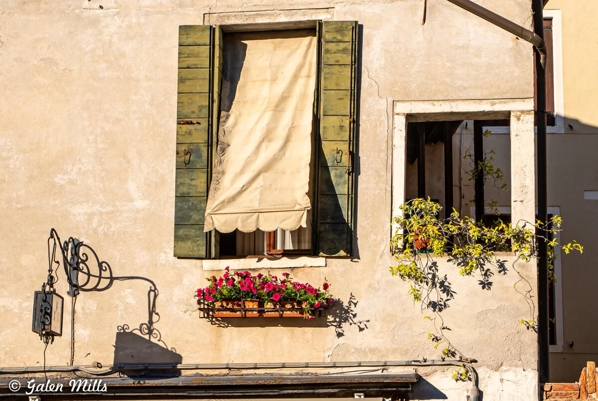 Facade of a rustic building with a window, green shutters, and a beige fabric shade. Below the window is a box of vibrant pink flowers. To the right, there's a partially open alcove with climbing greenery. Shadows and wiring are visible on the wall.