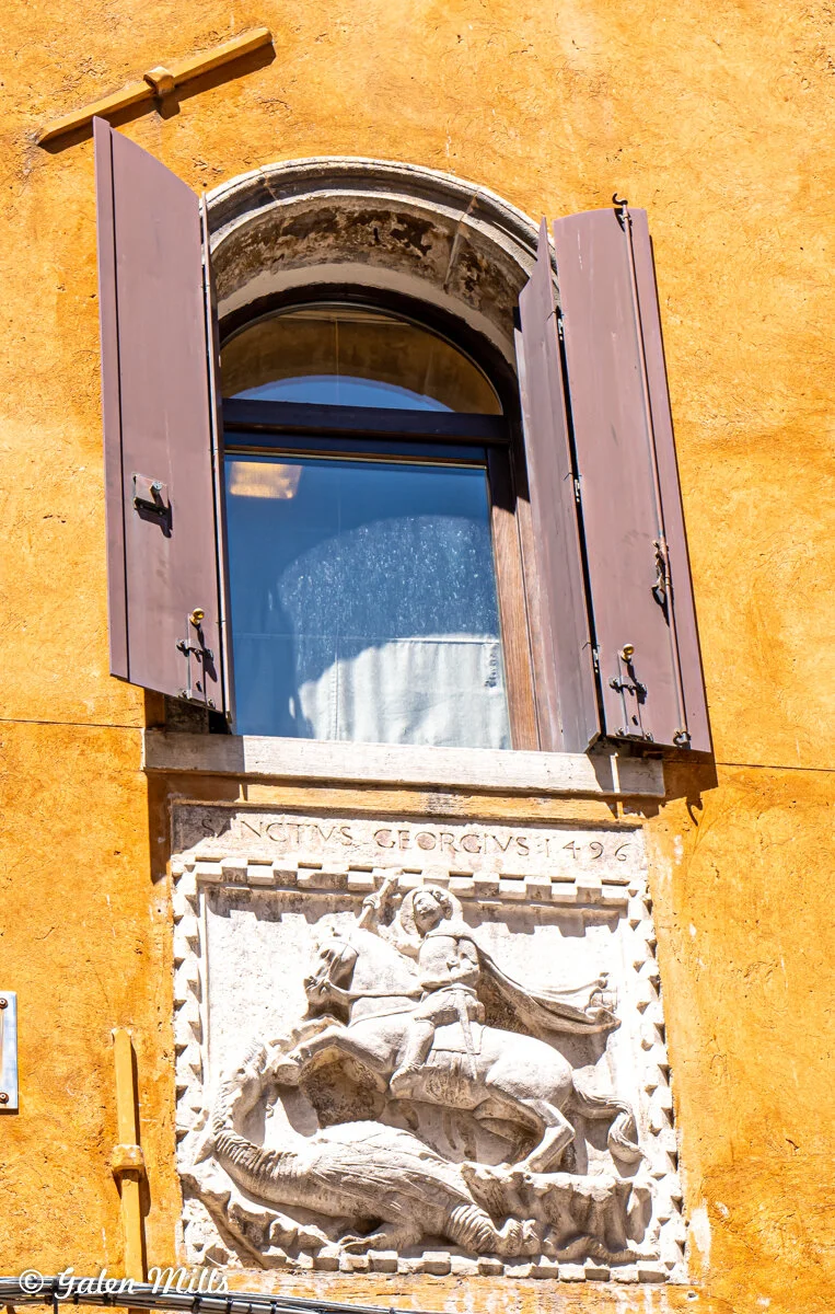 Arched window with brown shutters above a stone relief showing St. George slaying a dragon, dated 1496, set in a yellow wall.